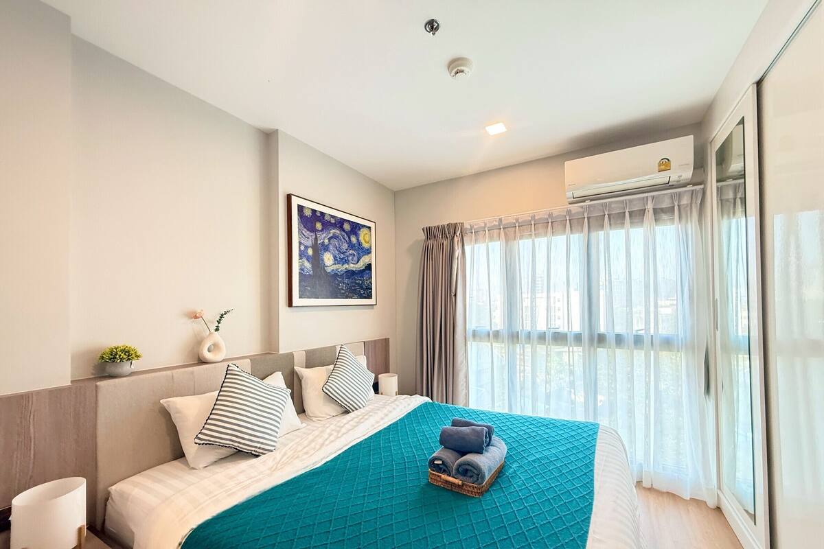 A modern hotel room with a large bed, a painting on the wall, and a view through sheer curtains.