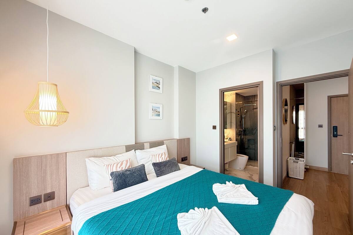 A modern hotel room with a large bed, a bedside table, a hanging light fixture, and a shower area visible through an open door.