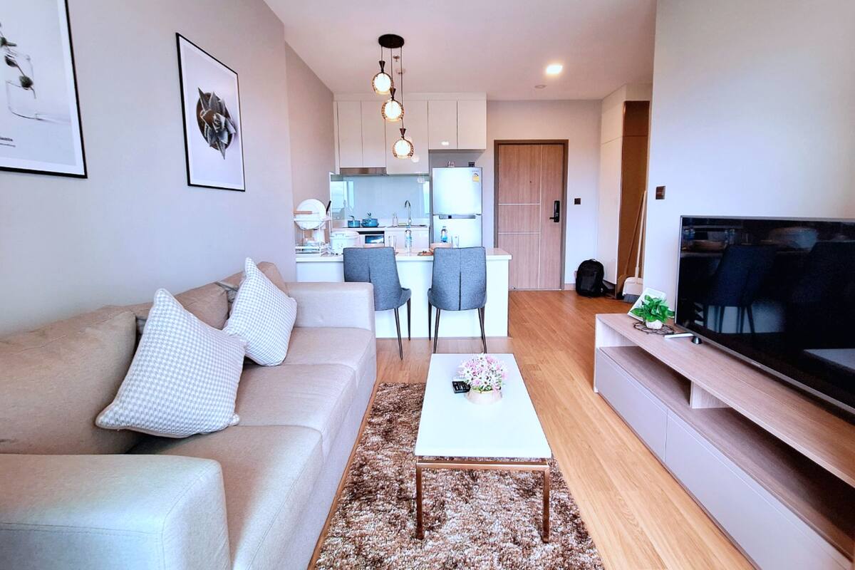 A modern living room with a sofa, coffee table, and TV stand. There is a kitchen in the background with a dining area.