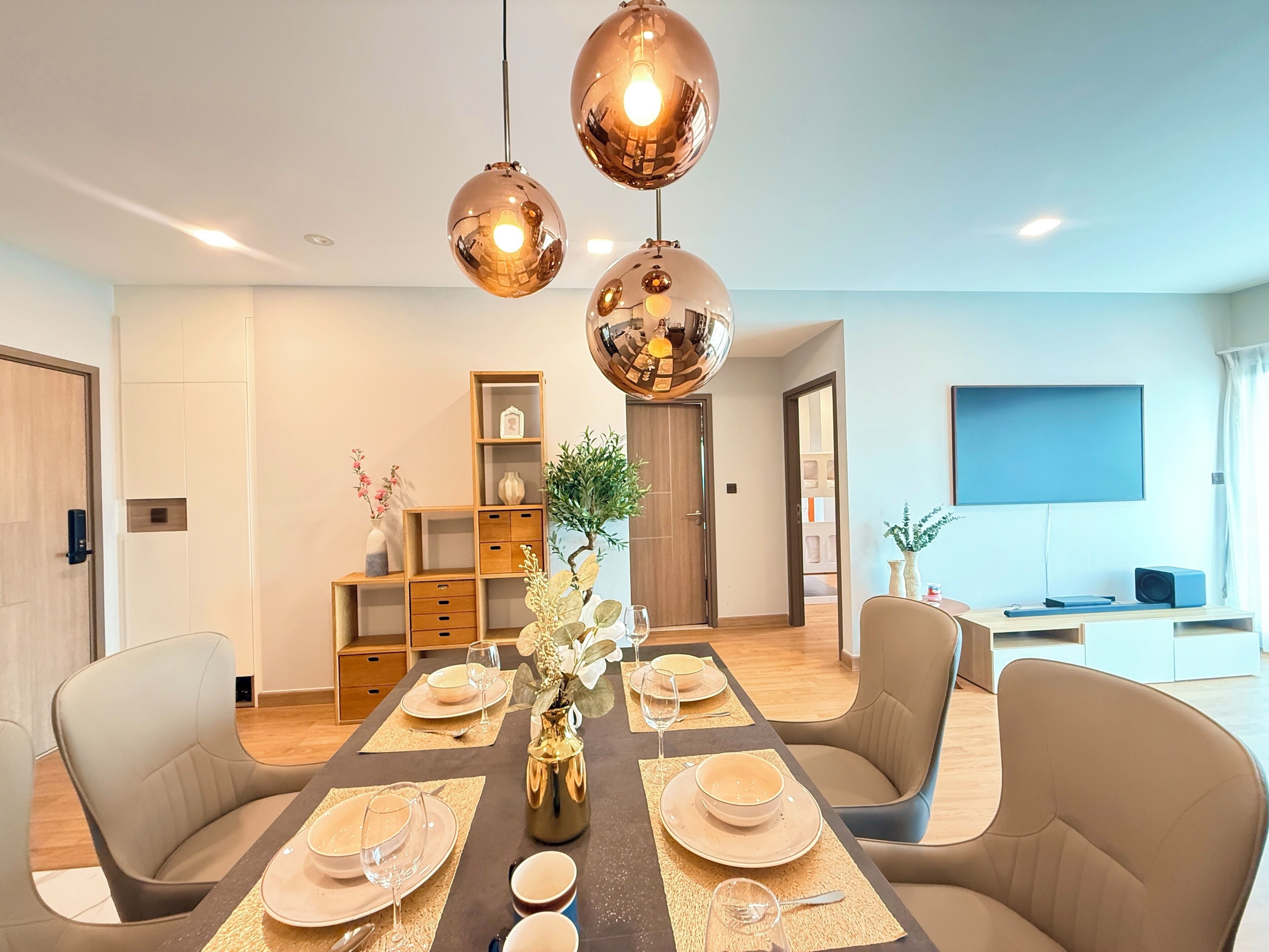 A dining area with a table set for four, two pendant lights, a bookshelf, a television, and a door leading to another room.