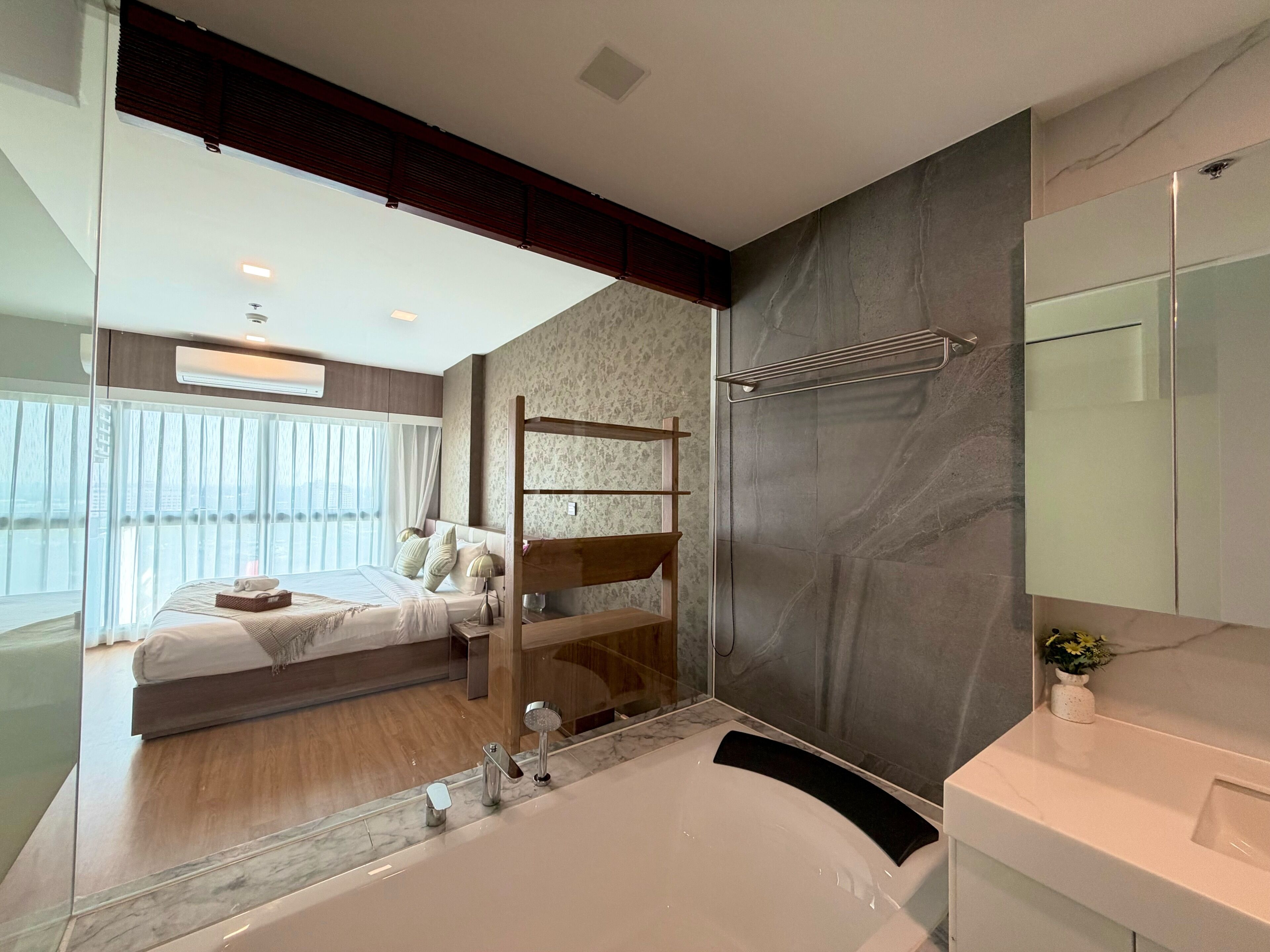 Une chambre à coucher moderne avec un lit, une tête de lit en bois, une grande fenêtre et une salle de bain équipée d’une baignoire et d’un miroir.