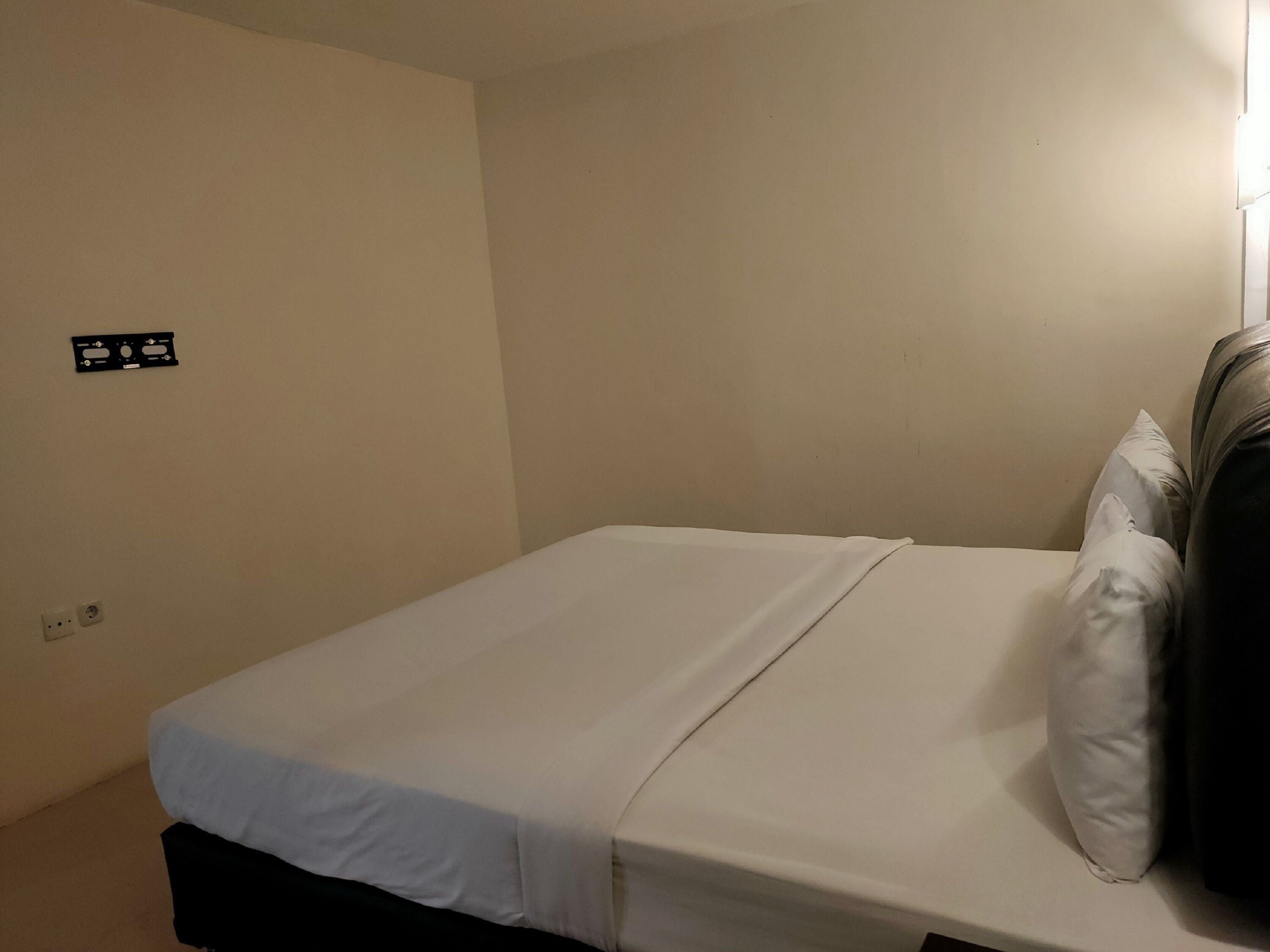 Una cama individual con ropa blanca en una habitación de paredes lisas y un enchufe a la izquierda.