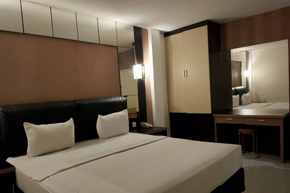 A modern hotel room with a large bed, a desk, a chair, a wardrobe, and a mirror.