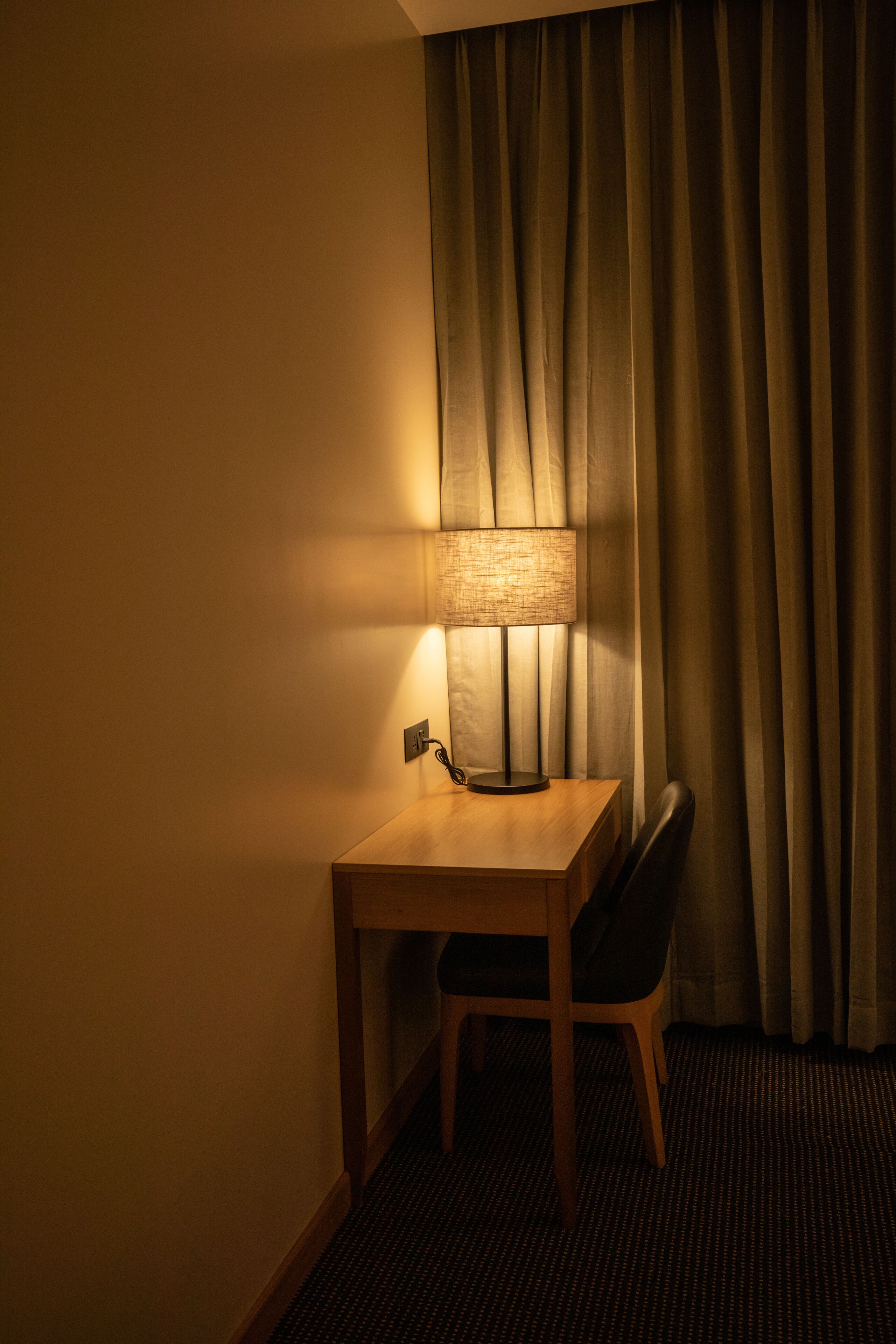 A hotel room with a wooden desk, a chair, a lamp, and a window with curtains.
