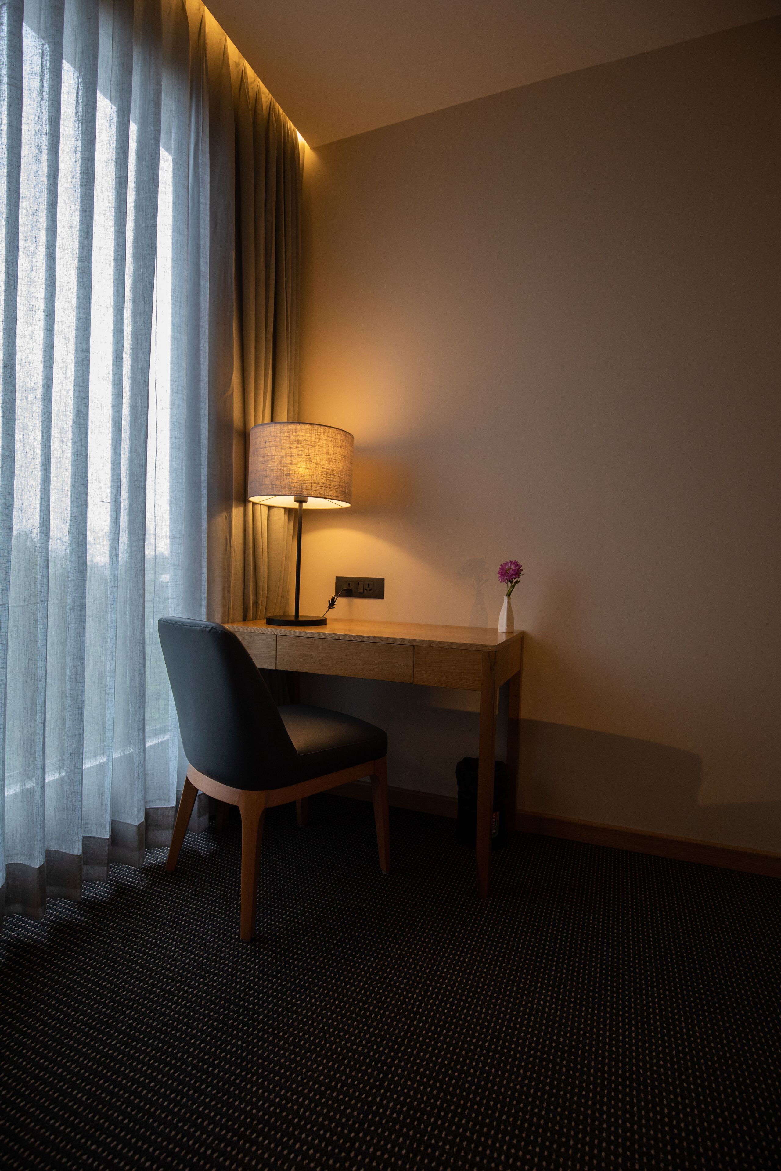 A hotel room with a desk, chair, lamp, and a vase with a flower.