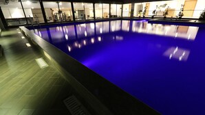 Una piscina interior con tonalidades azules, rodeada por un suelo de baldosas y un muro de cristal.