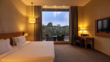 A hotel room with a bed, a desk, a chair, a television, and a balcony with a view of trees.