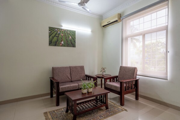 A living room with a sofa, two armchairs, a coffee table, and a small table with a plant. There is a painting on the wall, a ceiling fan, and a large window with blinds.
