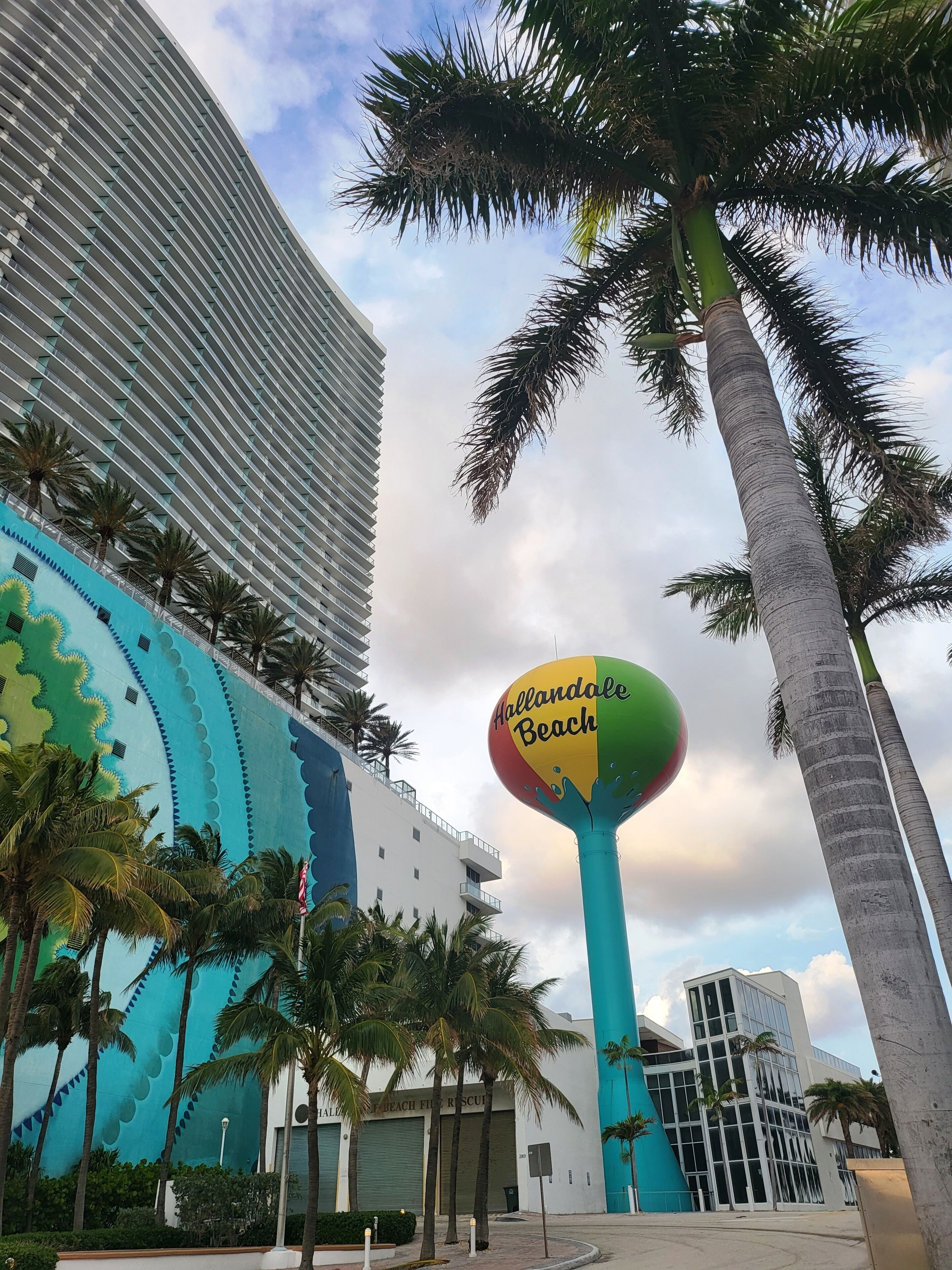 A modern building with a large mural, palm trees, and a beach-themed sign.