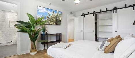 A bedroom with a bed, a ceiling fan, a TV, a plant, and a white brick wall.