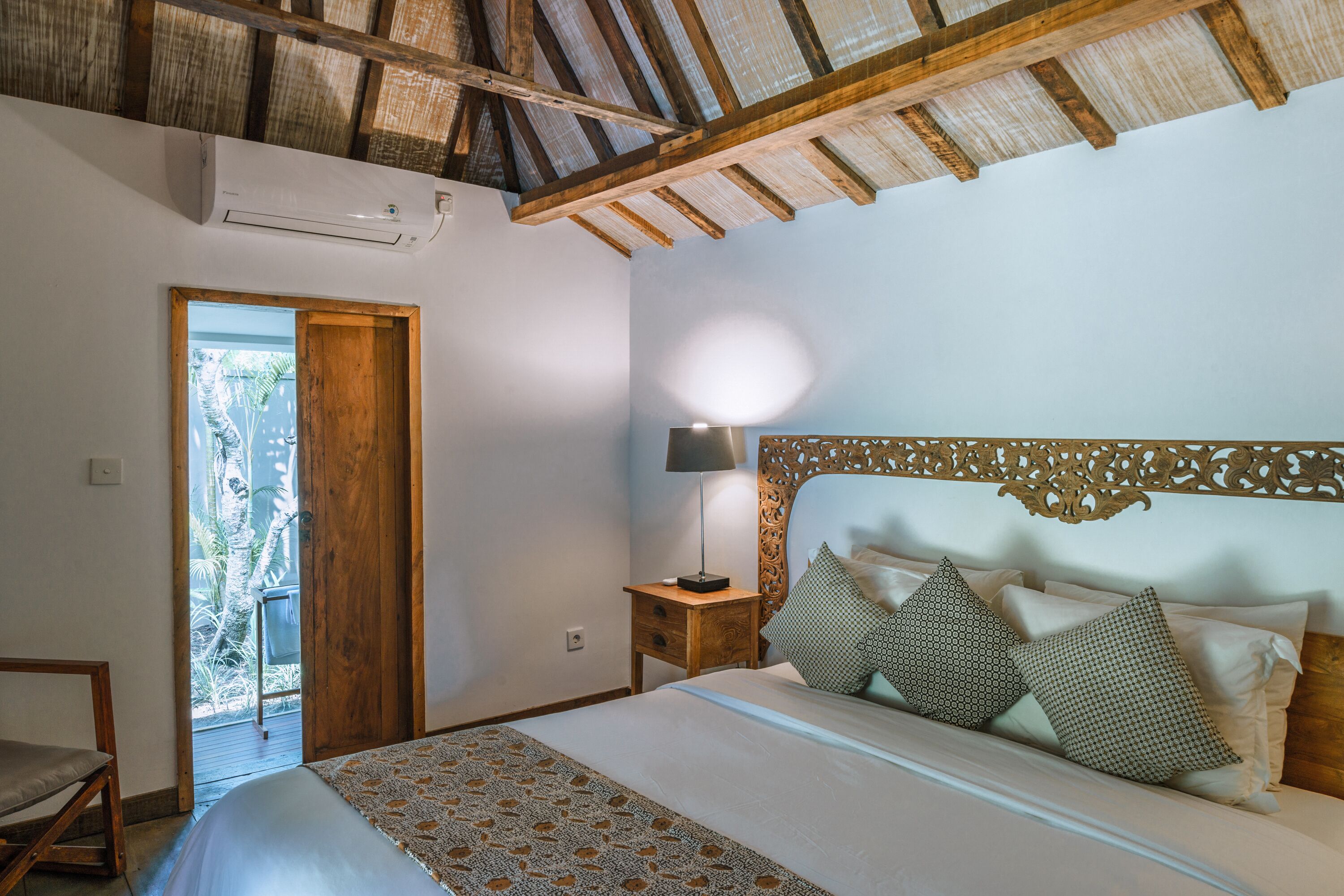 Ein Schlafzimmer mit Holzboden, einem Bett mit gemusterten Betttextilien, einem Nachttisch mit Lampe und Blick ins Freie durch eine Tür.