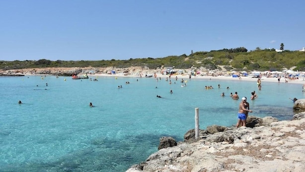 Una platja amb aigua cristal·lina, costa rocosa i persones nedant i prenent el sol.