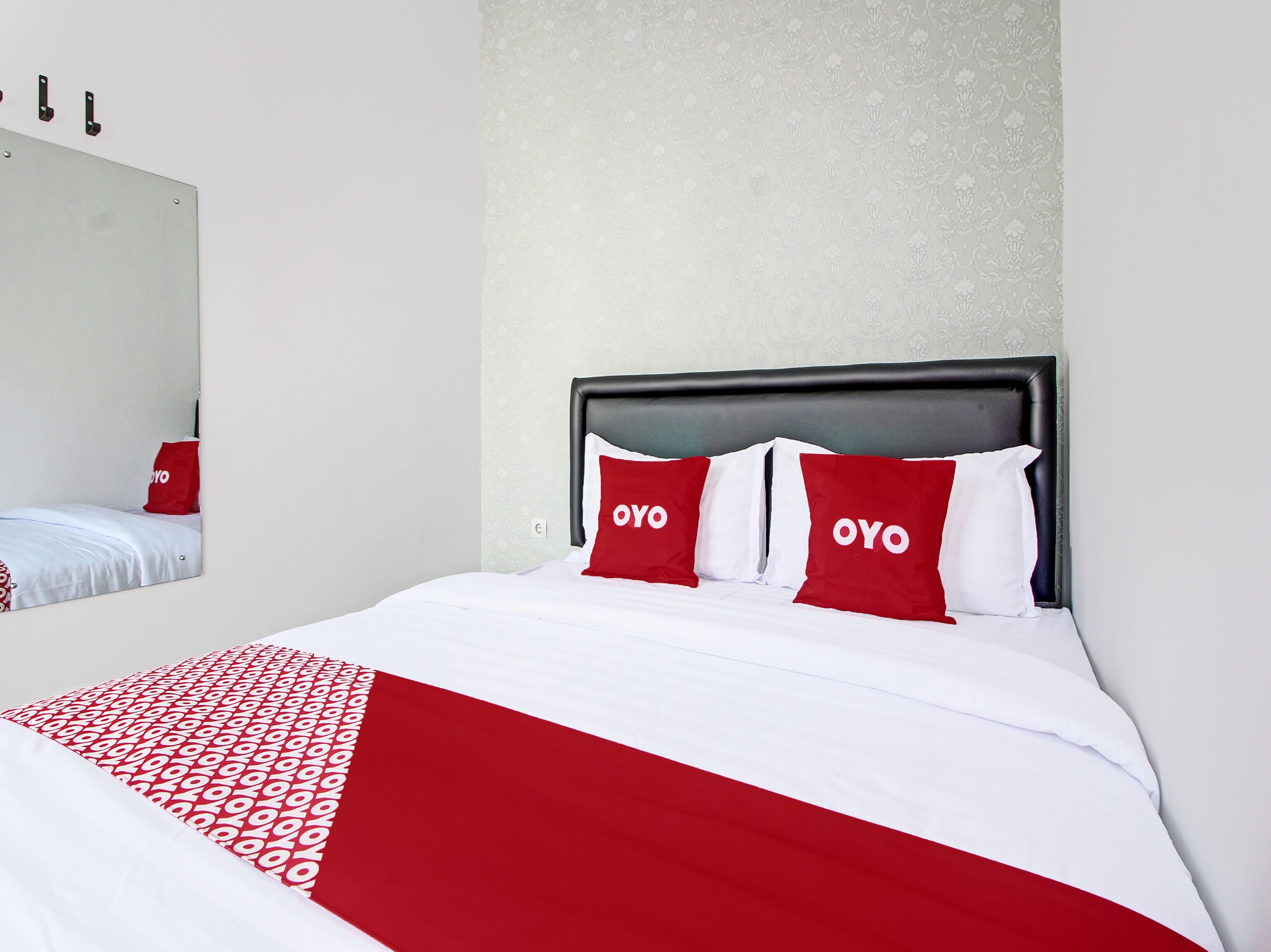 Een netjes opgemaakt bed met een rode en witte spreideken en kussens, een zwart hoofdbord en een spiegel aan de muur.