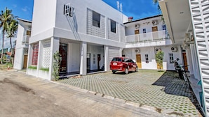 Un edificio blanco de dos pisos con un coche rojo estacionado en la entrada, rodeado de un área pavimentada con un diseño decorativo.