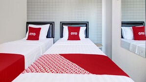 A hotel room with two beds, each with a black headboard and red and white bedding. The room has a tiled wall and a large mirror on one side.