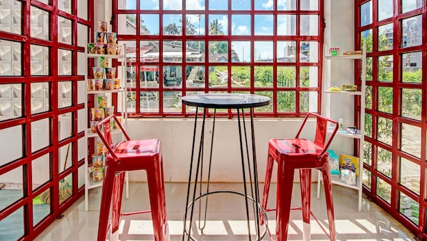 Una habitación con un ventanal grande, dos sillas rojas, una mesita y estanterías con diversos objetos.