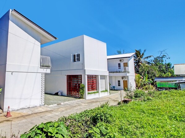Una casa moderna de dos pisos con un portón rojo, un balcón y un pequeño jardín.