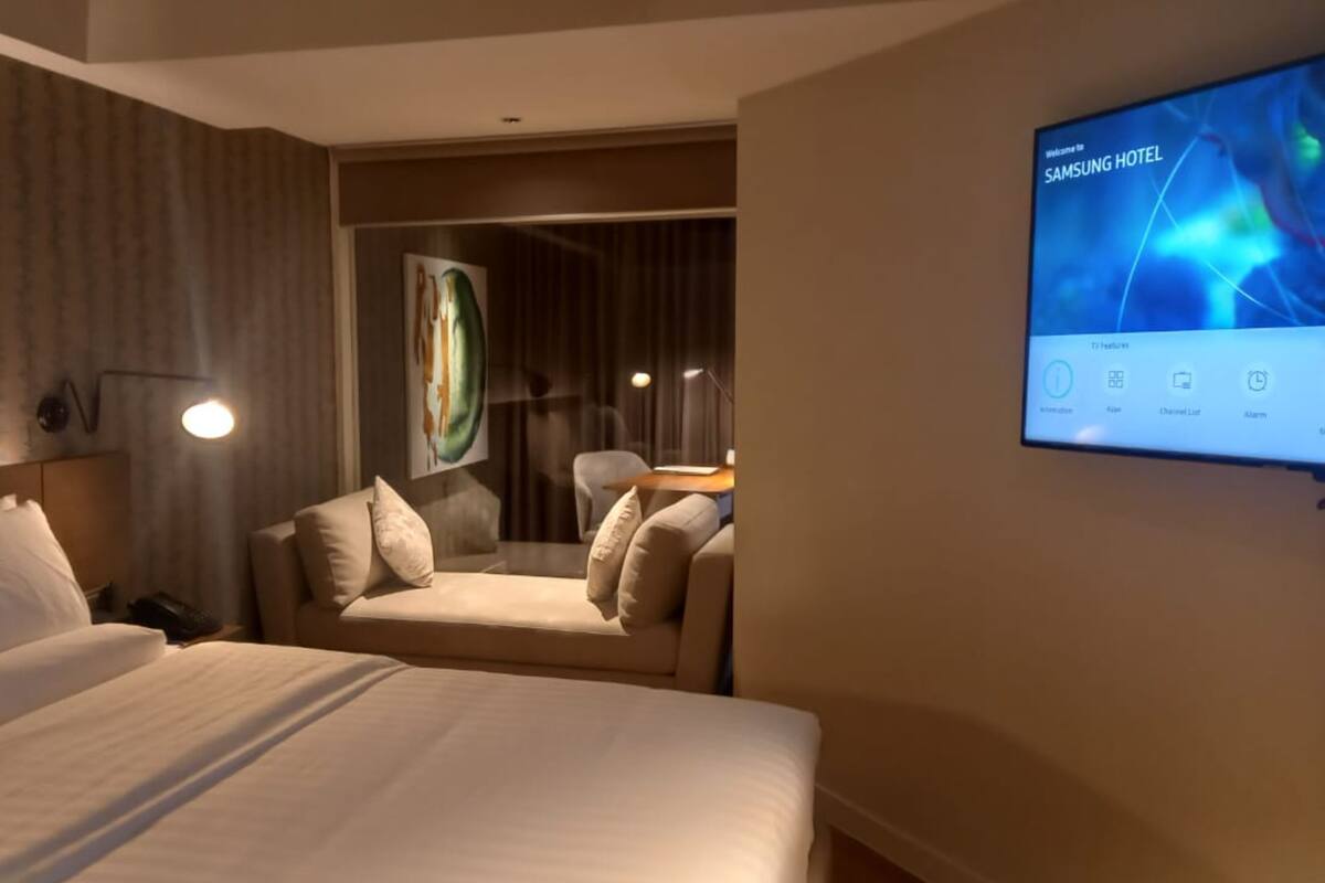 A hotel room with a bed, a sofa, a TV screen displaying 'Welcome to Samsung Hotel', and a painting on the wall.