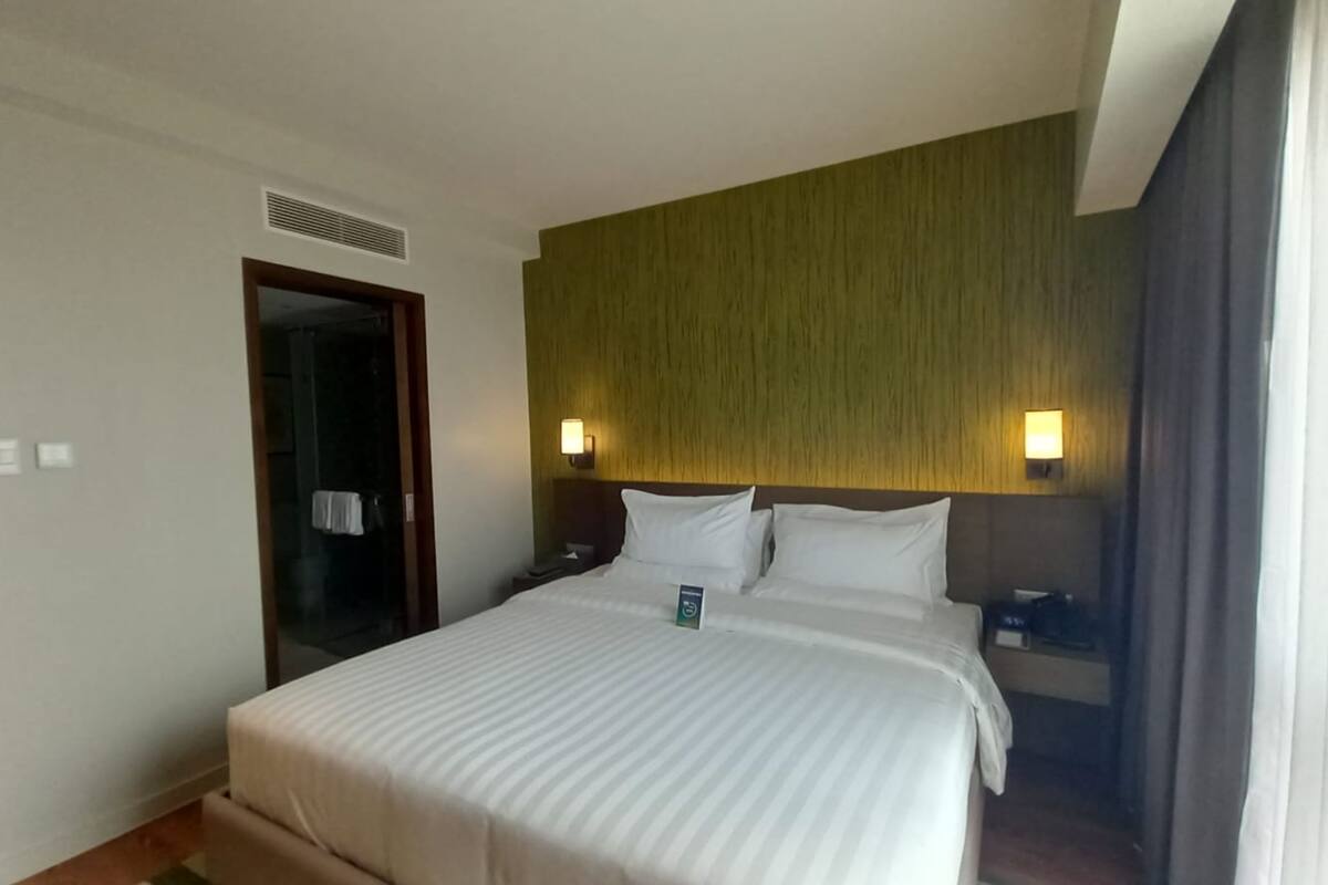 A hotel room with a large bed, a wooden headboard, two bedside tables, a nightstand, a wall-mounted light, and a door leading to a bathroom.