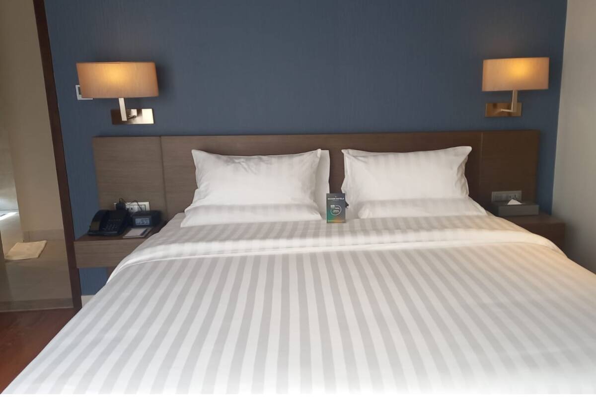 A hotel room with a large bed, two bedside lamps, a nightstand with a phone and a box, and a wooden headboard.