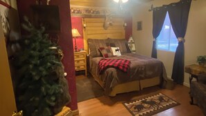 A cozy bedroom with a wooden ceiling, a bed with a red and black checkered blanket, a Christmas tree, and a window with curtains.
