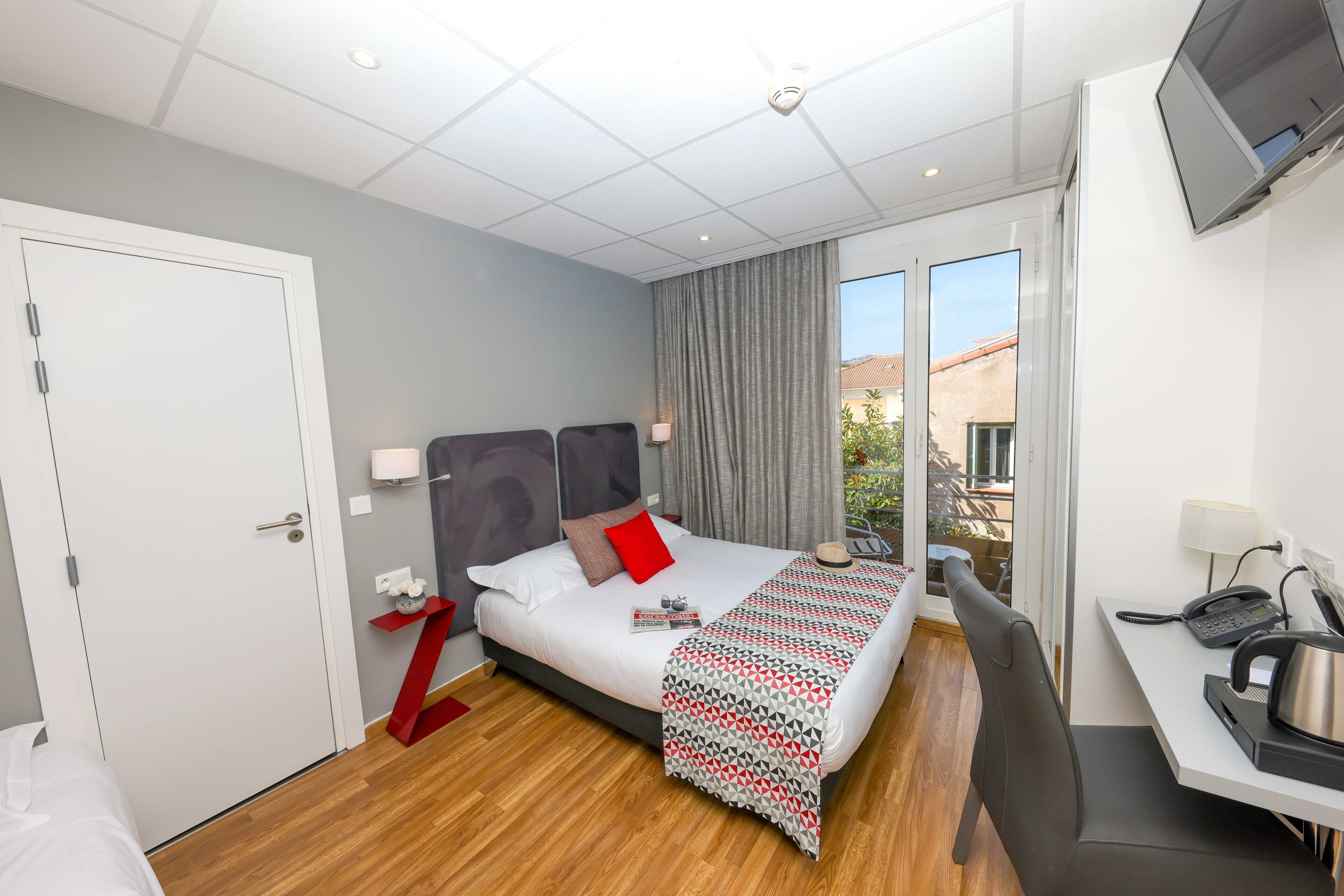 Une chambre d’hôtel équipée d’un lit, d’un bureau, d’une chaise, d’une télévision et d’une fenêtre donnant sur des immeubles.