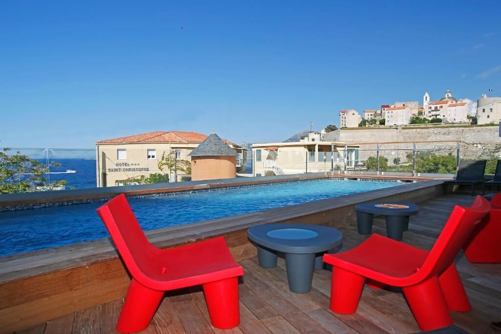 A rooftop pool area with red chairs and tables, overlooking a coastal town.