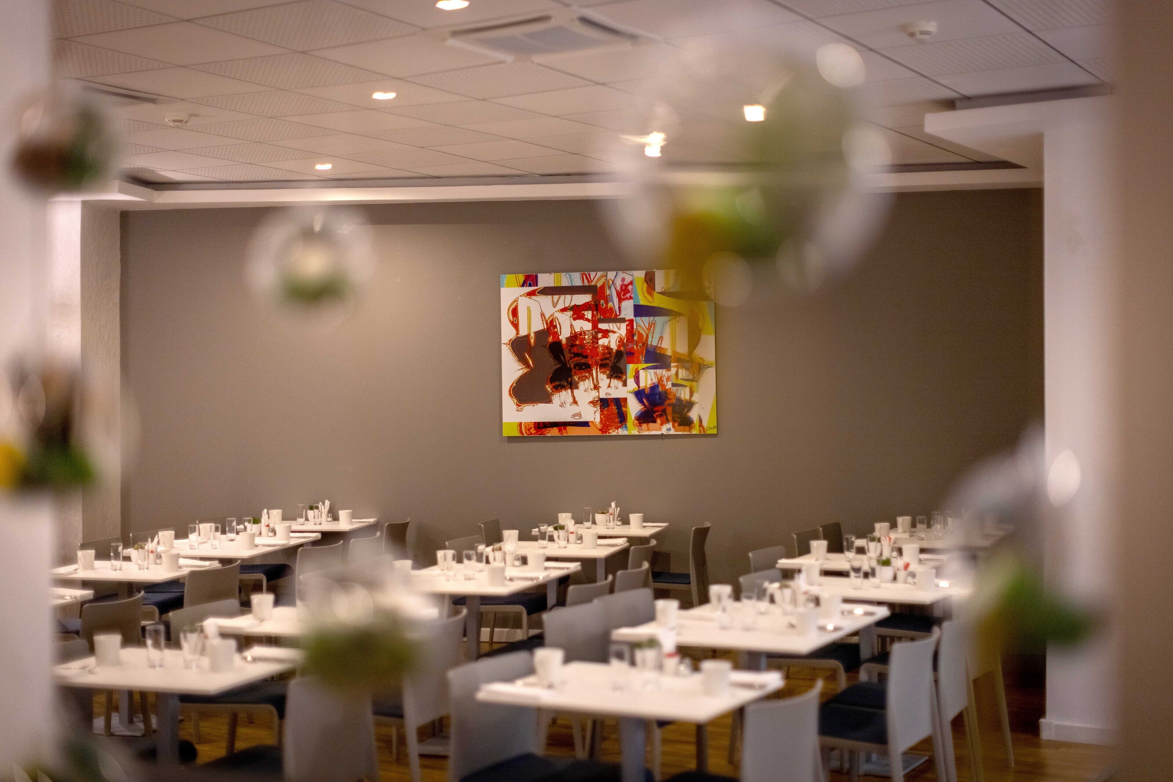 A dining area with tables set for service, a large abstract painting on the wall, and a ceiling with recessed lighting.
