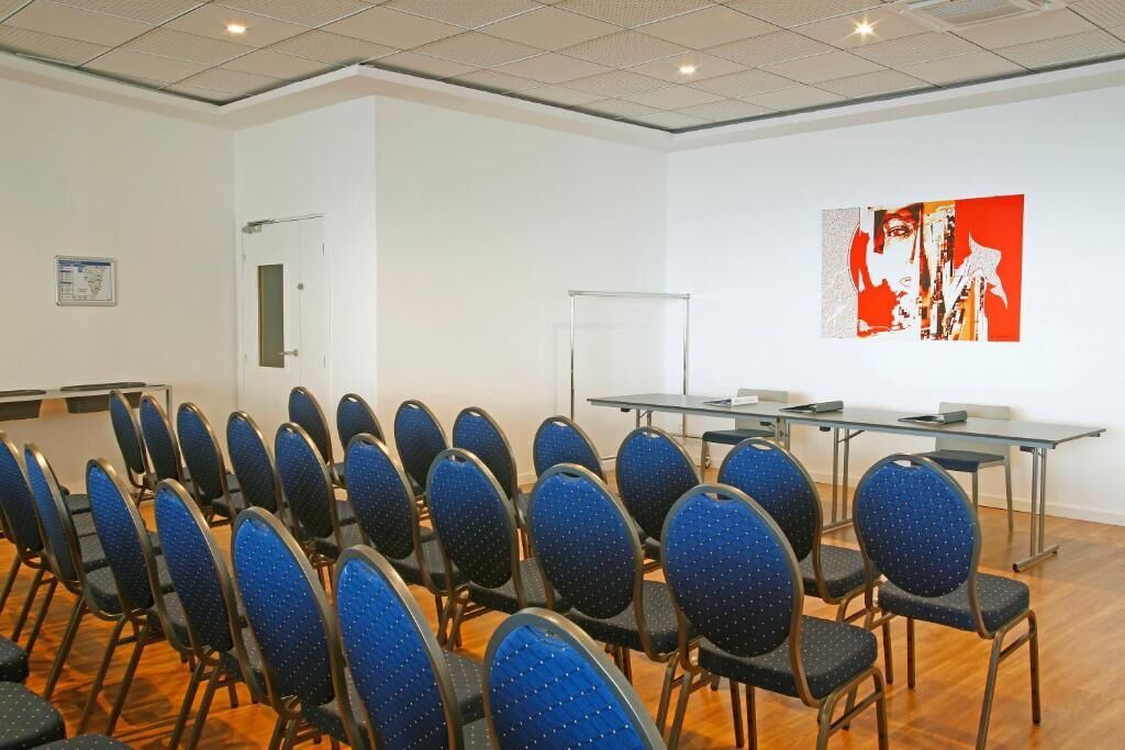 Une salle de conférence avec des rangées de chaises bleues, un tableau blanc et un grand tableau abstrait accroché au mur.