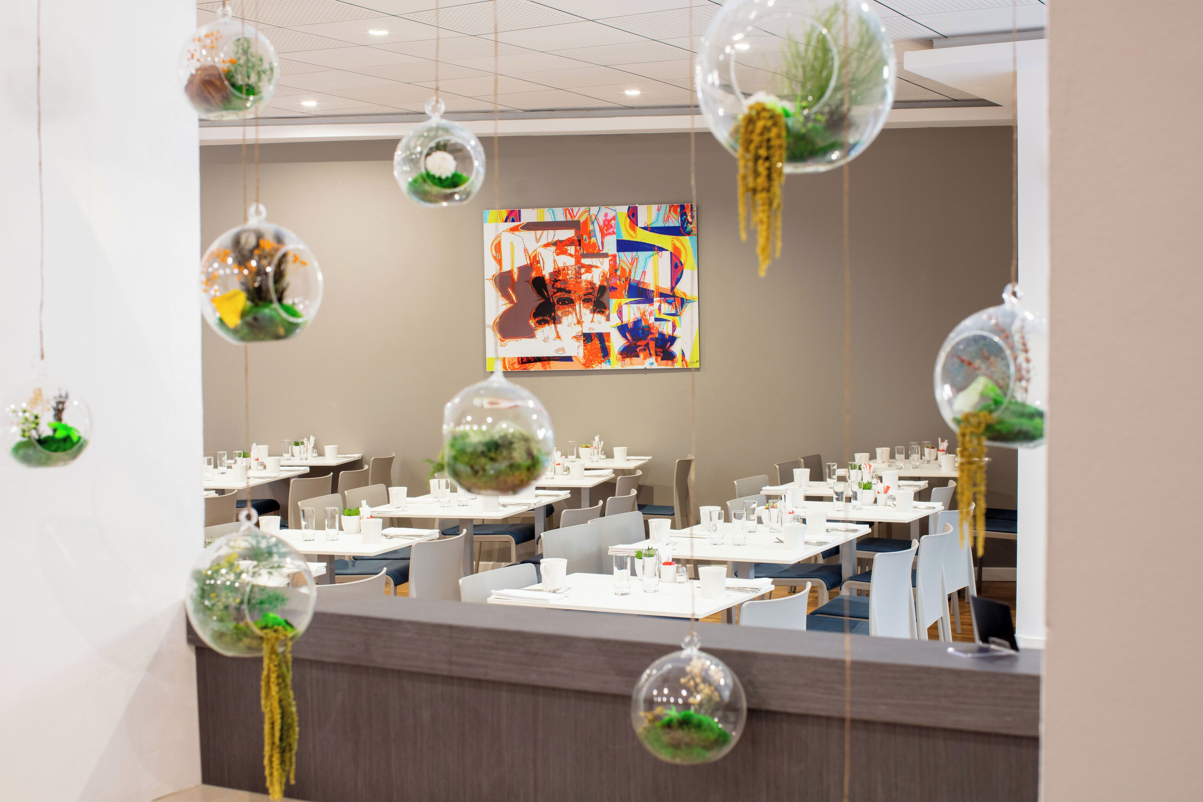 Un espace de restauration moderne avec des tables dressées, une grande peinture abstraite au mur et des sphères décoratives suspendues, ornées de verdure.