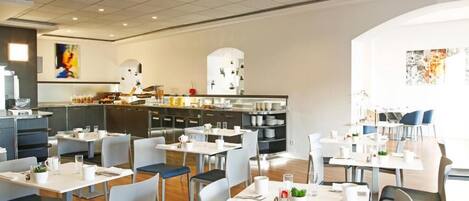 Un comedor moderno con mesas y sillas blancas, una cocina al fondo y obras de arte en las paredes.