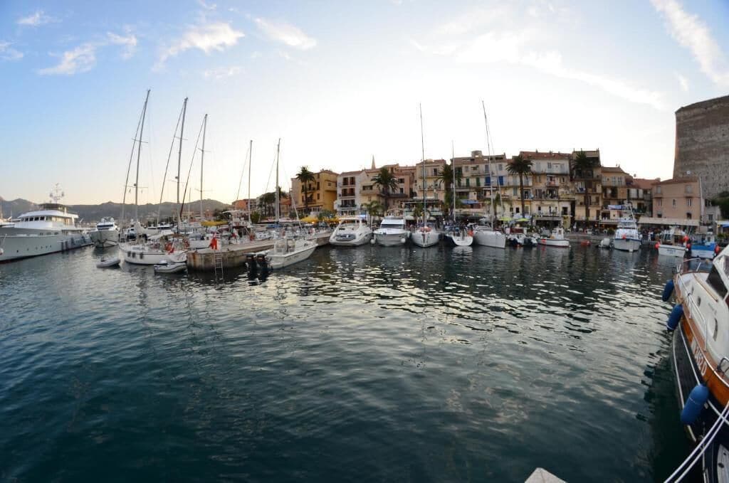 A marina with multiple boats docked, a waterfront promenade, and buildings with balconies overlooking the water.