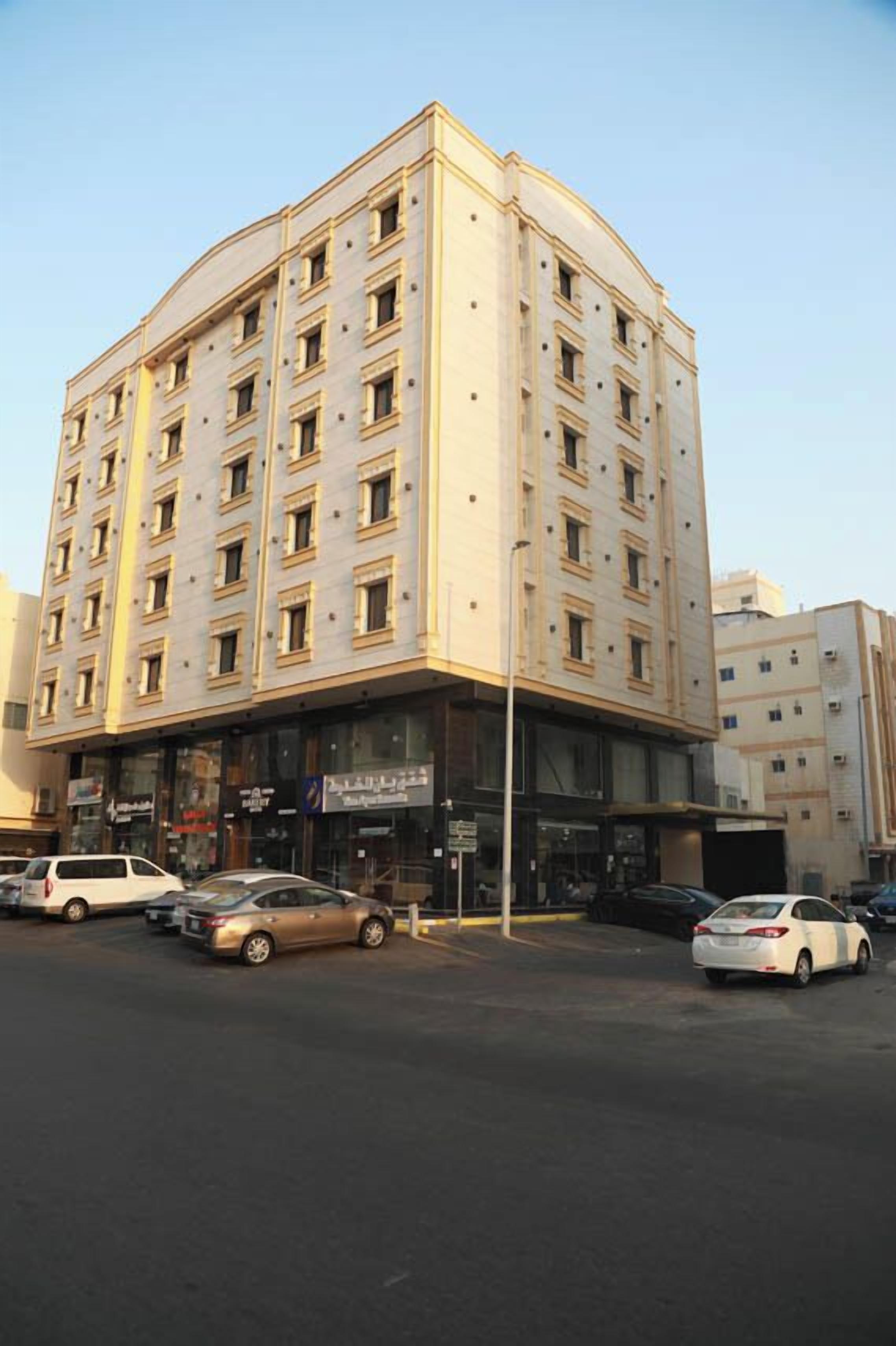 A multi-story hotel with a corner entrance, several ground-floor shops, and a parking lot with cars.