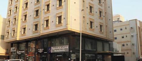 A multi-story hotel with a corner entrance, several ground-floor shops, and a parking lot with cars.