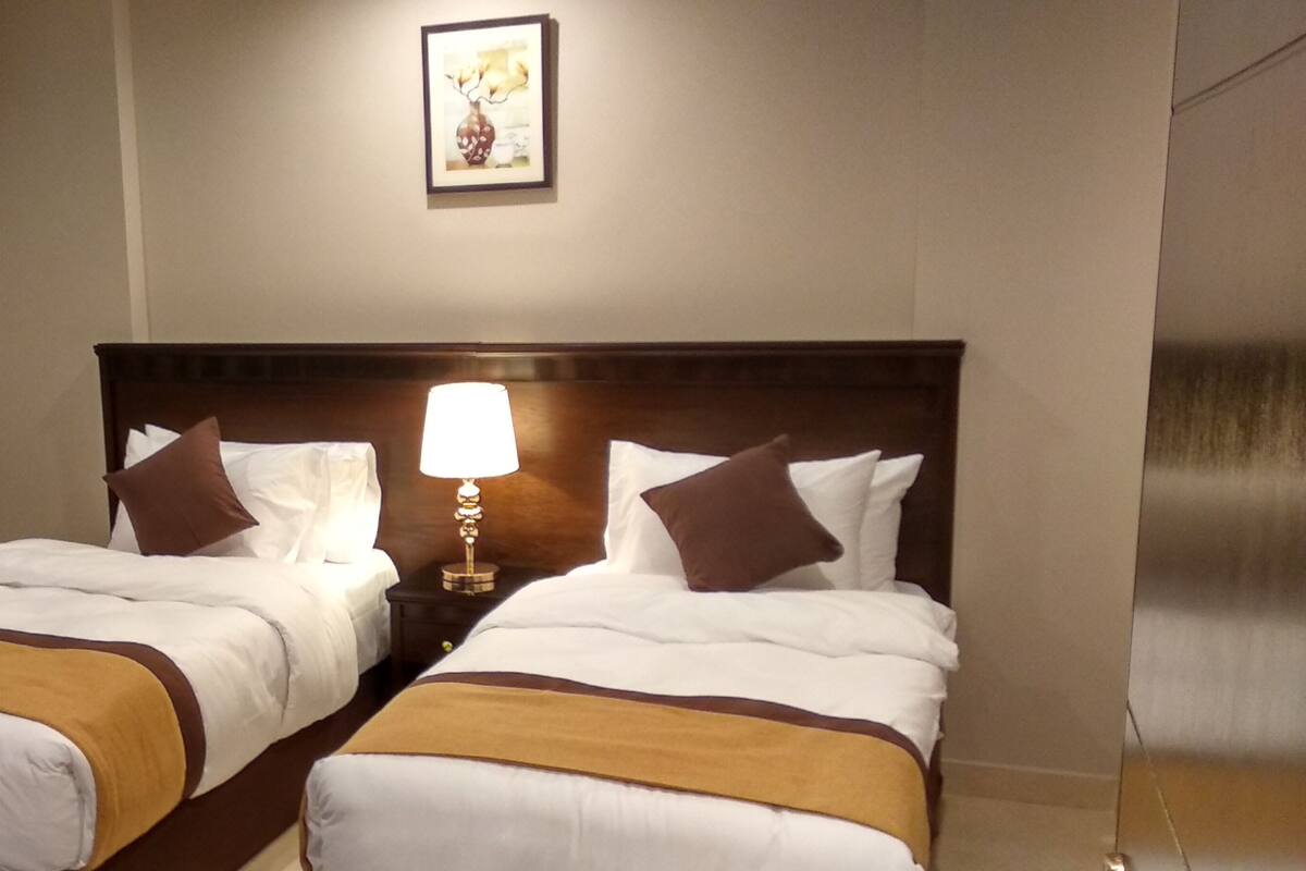 Ein Hotelzimmer mit zwei Betten, einem Nachttisch mit Lampe, einer wandmontierten Klimaanlage und einem gerahmten Bild über den Betten.