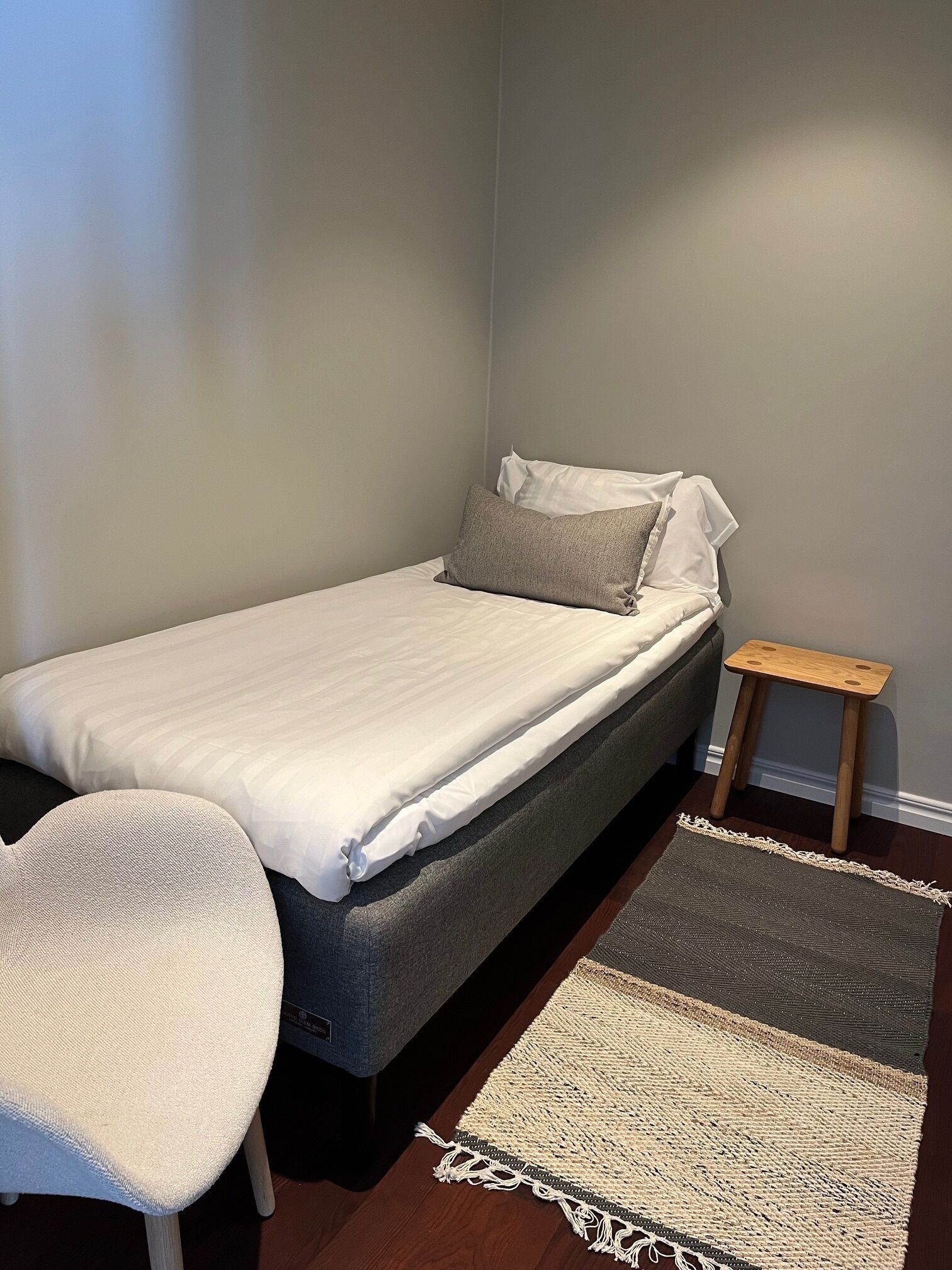 Una cama individual con colchón gris y ropa de cama blanca, una pequeña mesita de noche de madera, un tapete tejido y una silla blanca.