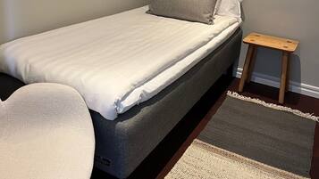 A single bed with a grey mattress and white bedding, a small wooden nightstand, a woven area rug, and a white chair.