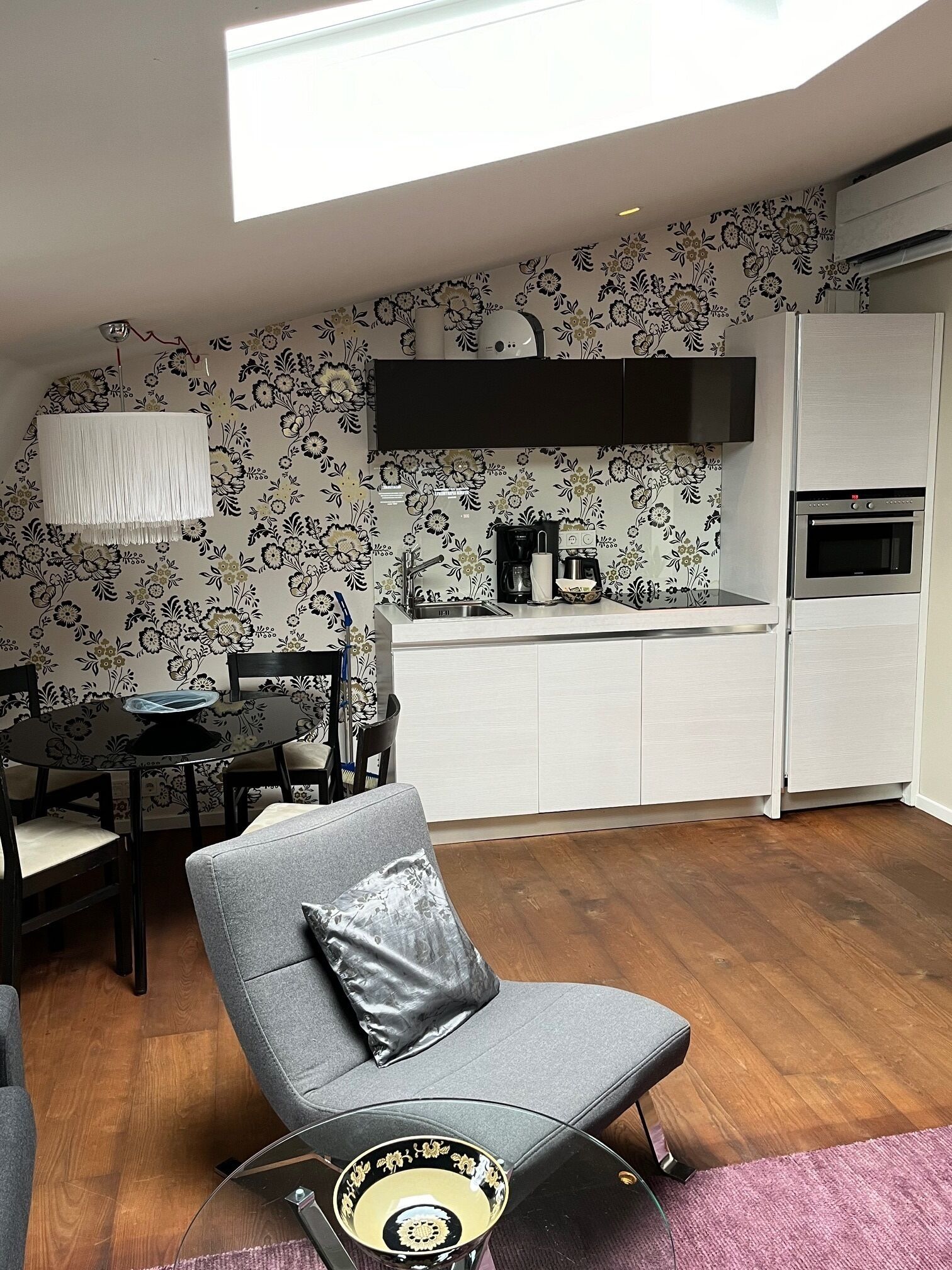 A modern kitchen with a dining area, featuring a black and white floral wallpaper, a built-in oven, and a dining table with chairs.