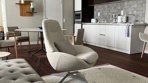 A modern living room with a tufted sofa, a round coffee table, and a dining area with a small table and chairs. A kitchen with a black range hood and white cabinets is visible in the background. - DA Hotel Apartments (Gothenburg)
