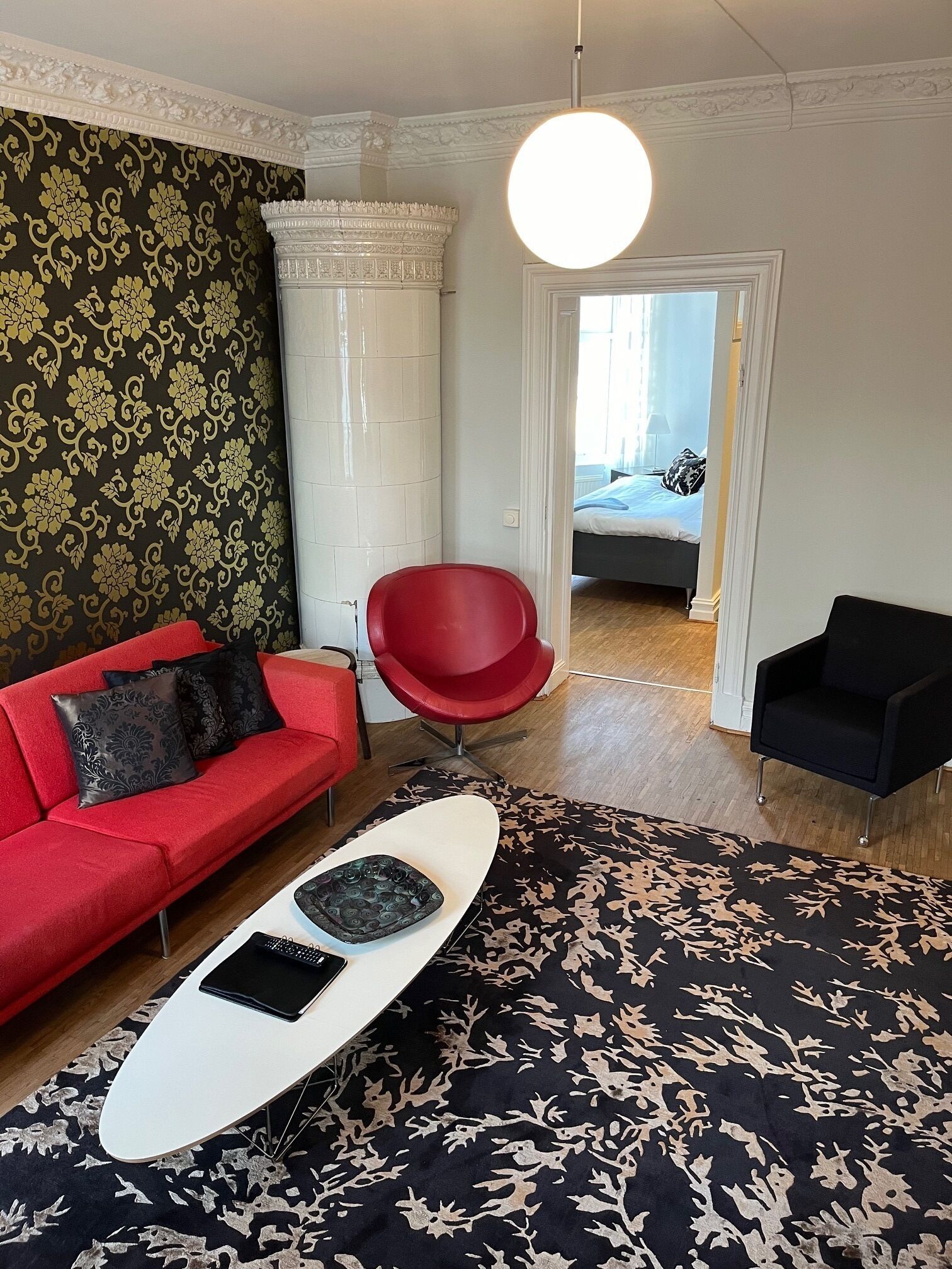 Una sala de estar moderna con un sofá rojo, una mesa de centro blanca y un tapete estampado. Hay una silla roja y una silla negra en la habitación.