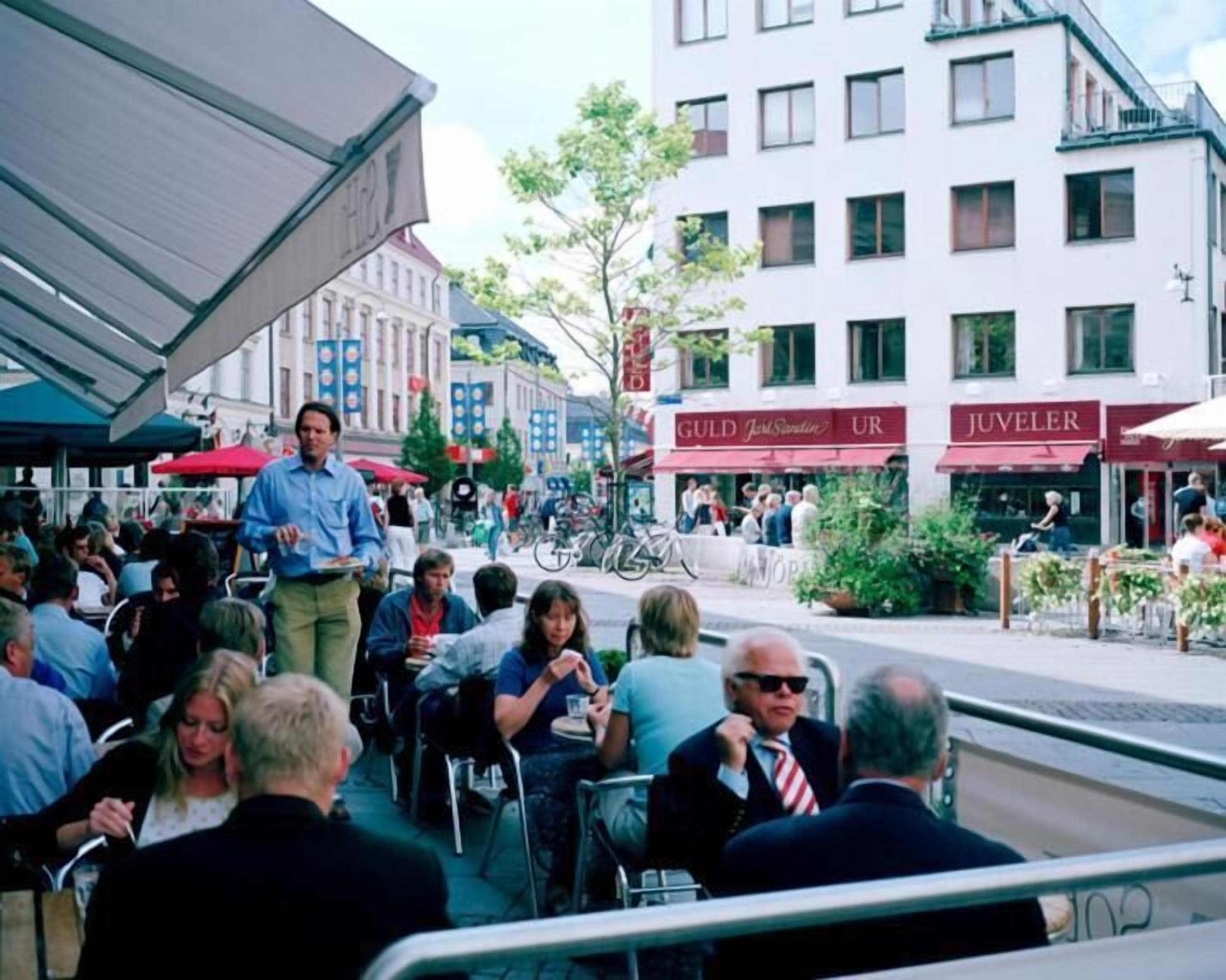 An outdoor café with tables and chairs, surrounded by buildings with visible signage.