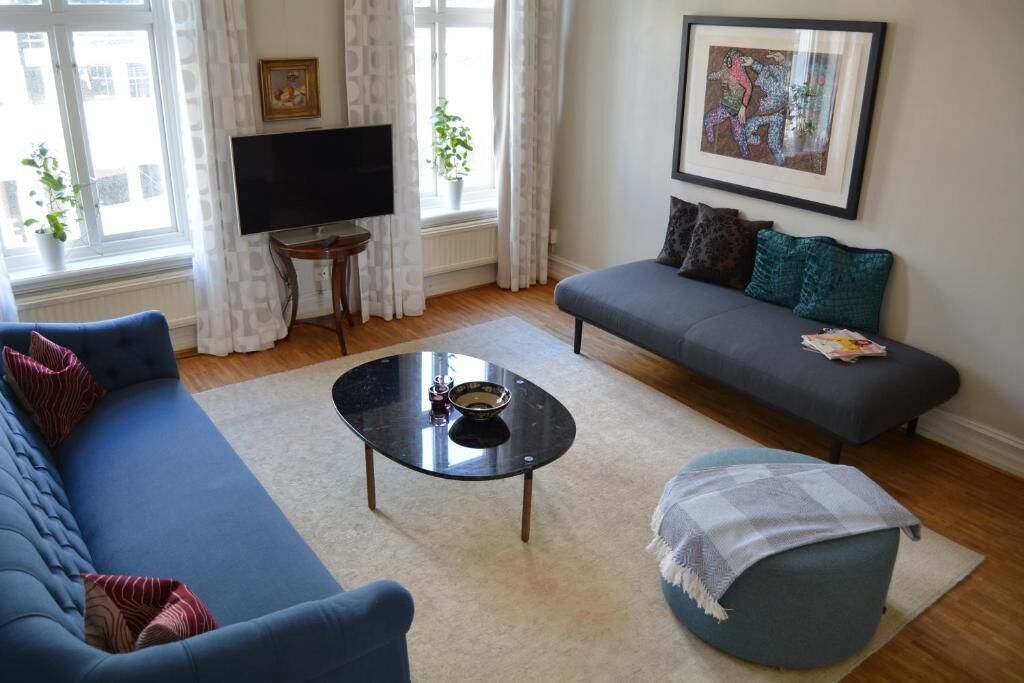 Ein Wohnzimmer mit blauem Sofa, grauem Sofa, rundem Couchtisch und blauem Hocker. Ein Fernseher steht auf einem Schrank, an der Wand hängt ein gerahmtes Bild und es gibt ein Fenster mit Vorhängen.
