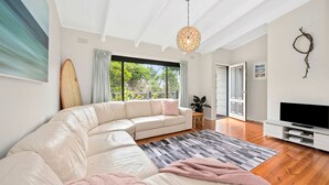 A modern living room with a large L-shaped sectional sofa, a flat-screen TV, and a large window offering a view of greenery. - Three Beaches (Anglesea)