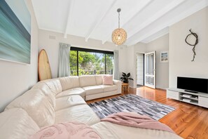 A modern living room with a large L-shaped sectional sofa, a flat-screen TV, and a large window offering a view of greenery. - Three Beaches (Anglesea)