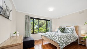 Quarto com cama de madeira, mesa de cabeceira, janela com cortinas, pequena mesa com uma planta e obra de arte na parede.