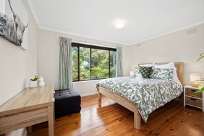 A bedroom with a wooden bed, a bedside table, a nightstand, a window with curtains, a small table with a plant, and a wall-mounted artwork. - Three Beaches (Anglesea)