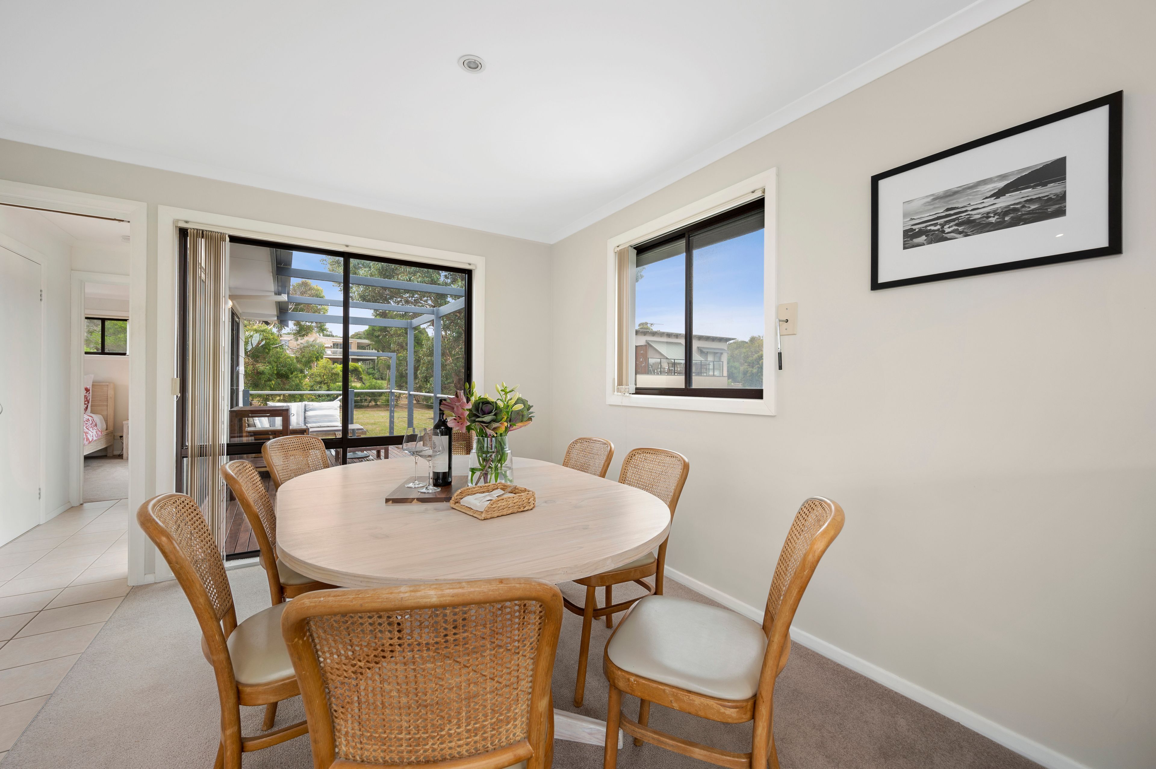 A dining area with a round table, six wicker chairs, a vase with flowers, and a framed picture on the wall.