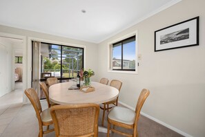 A dining area with a round table, six wicker chairs, a vase with flowers, and a framed picture on the wall. - Three Beaches (Anglesea)