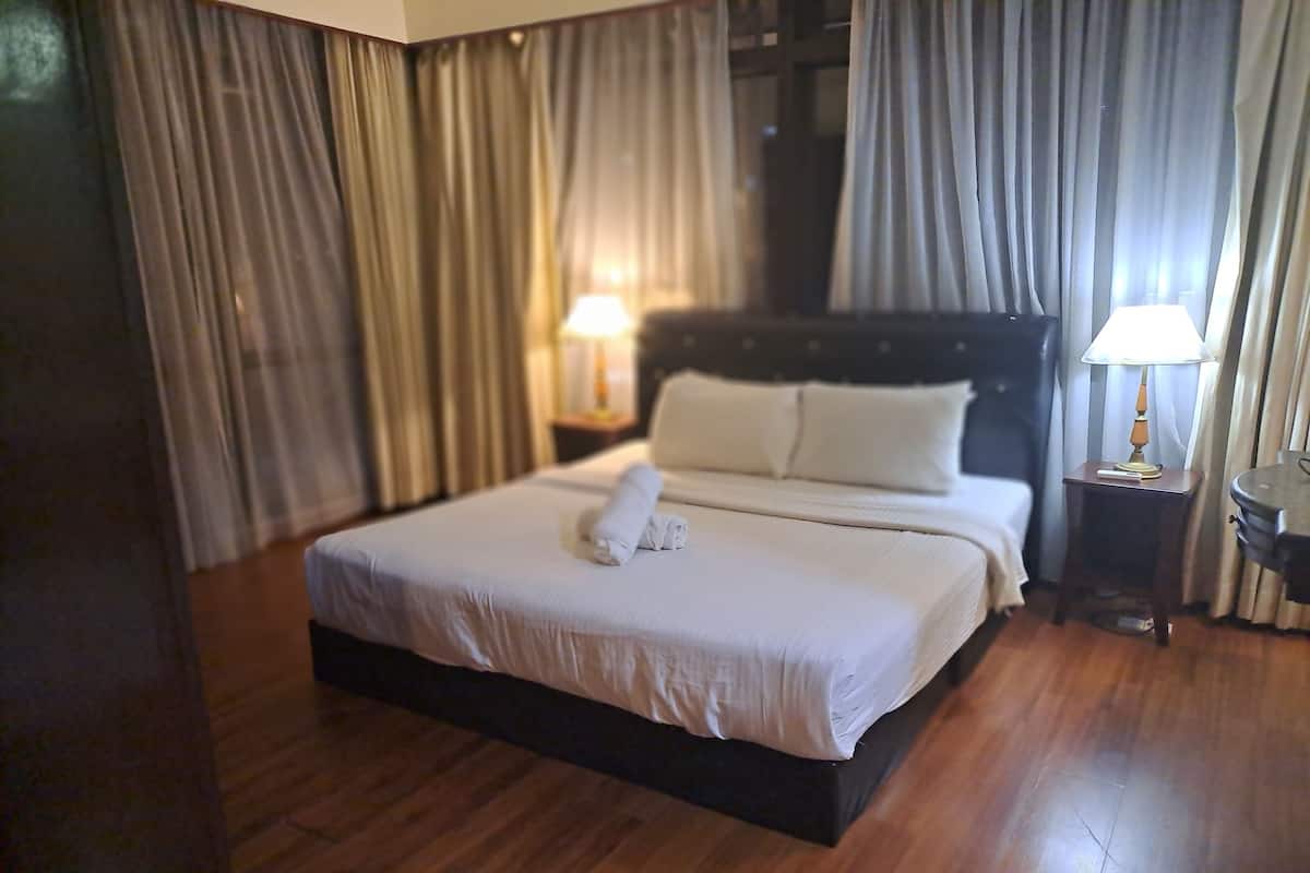 A neatly made bed with white linens and a rolled towel, a bedside table with a lamp, and a wooden floor.