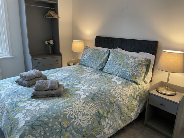 Una cama bien hecha con ropa de cama floral, dos lámparas de noche y un juego de toallas sobre la cama.